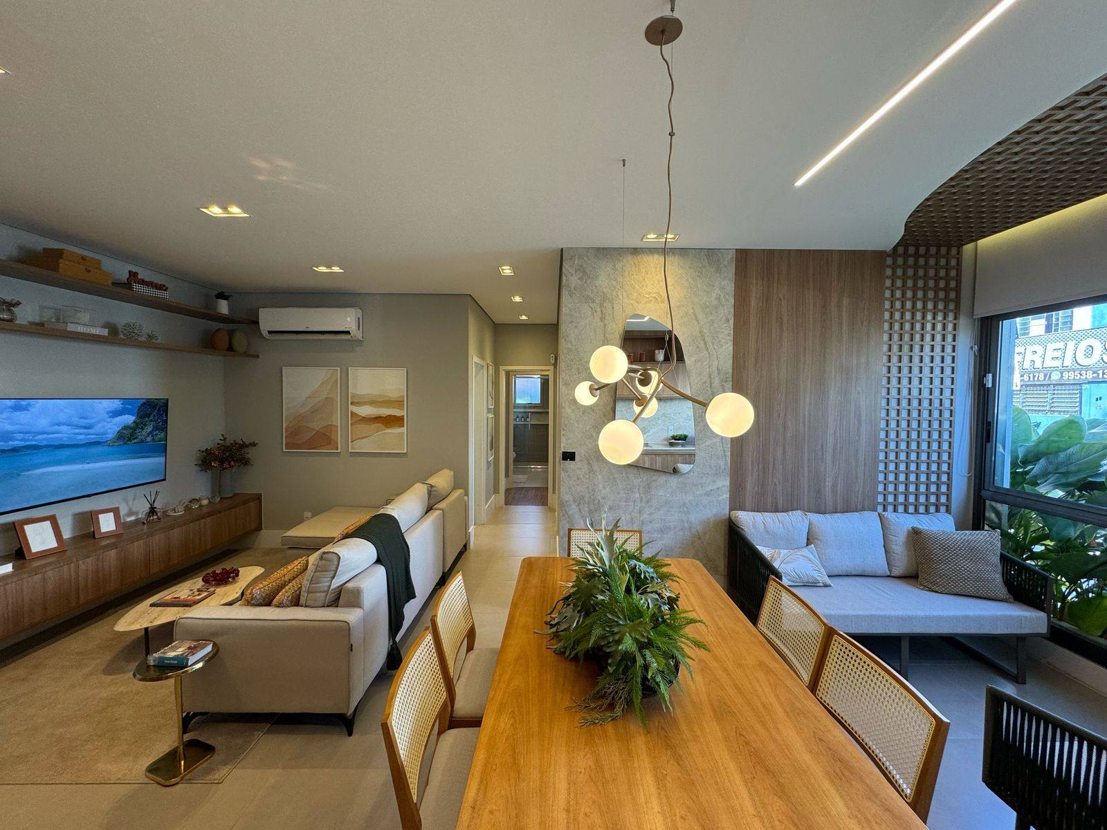 Sala de estar do decorado com painel de TV, luminária pendente e mesa de jantar de madeira junto à varanda