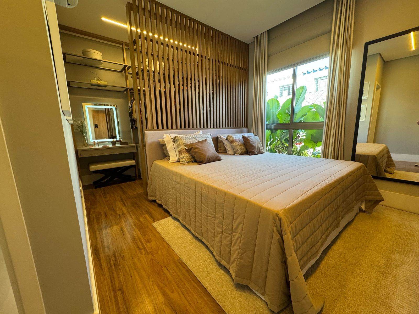 Suíte do apartamento decorado com cama de casal, painel ripado de madeira, penteadeira com espelho iluminado e janela ampla