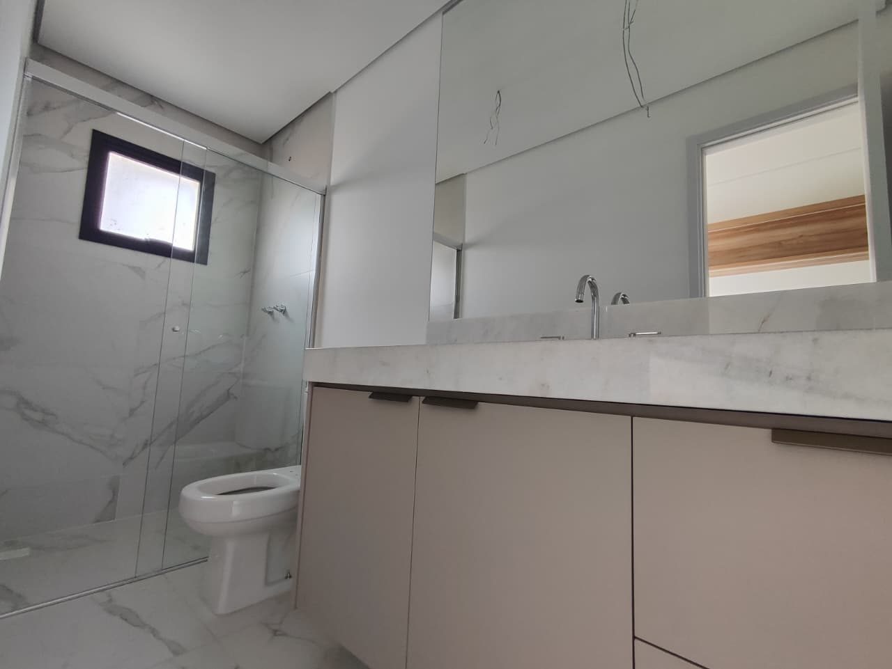 Foto real de um banheiro do apartamento 44, com box de vidro, revestimento claro com veios, bancada clara e gabinete claro