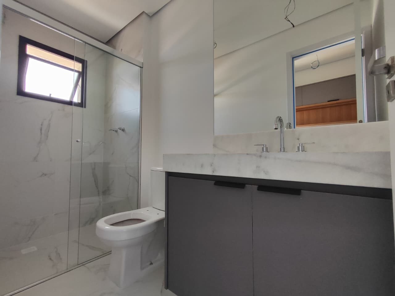 Foto real de um banheiro do apartamento 33, com box de vidro, revestimento claro com veios, bancada clara e gabinete cinza