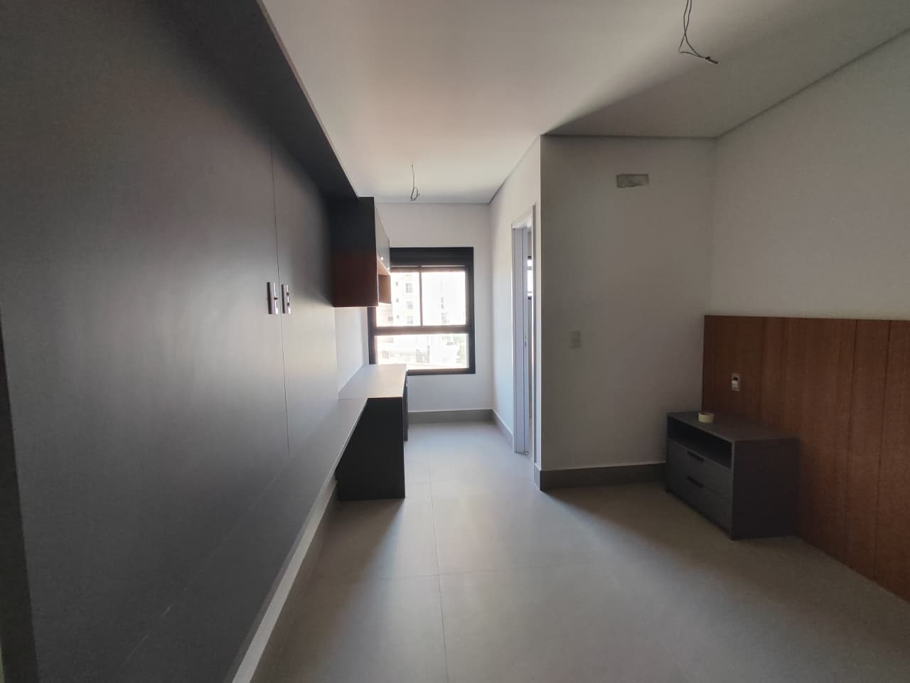 Foto real de uma suíte do apartamento 33, com painel cinza de armários, bancada sob a janela e cabeceira de madeira